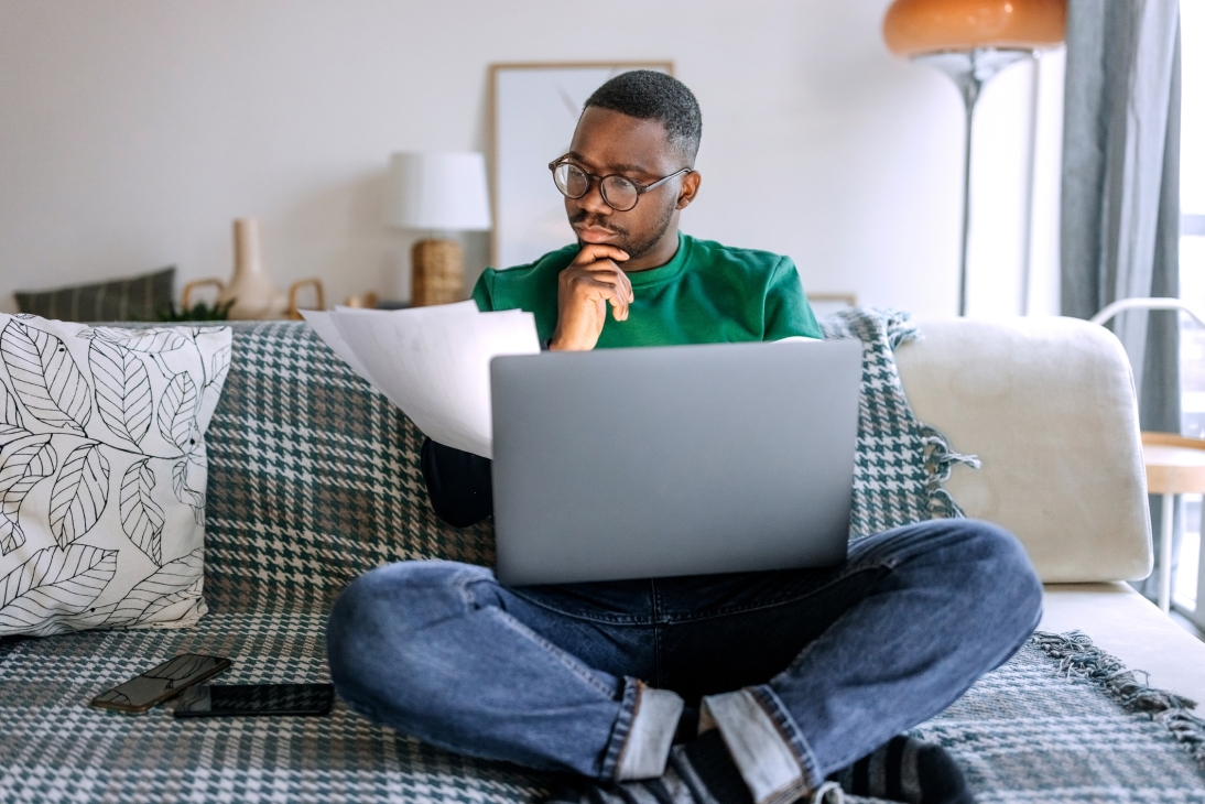 Jeune homme concentré, vêtu de façon décontractée, examinant la paperasse tout en travaillant depuis le salon, il est assis sur le canapé avec un ordinateur portable sur ses genoux
