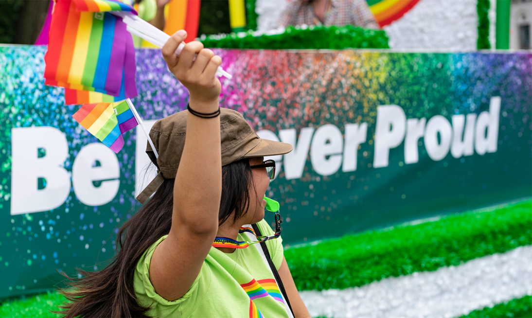 Woman standing and waving a pride flag while wearing a green TD shirt.