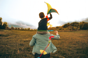 Father with his young son on his shoulders. The son is holding a kite as they run through a field.