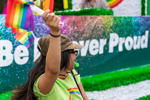 Woman standing and waving a pride flag while wearing a green TD shirt.